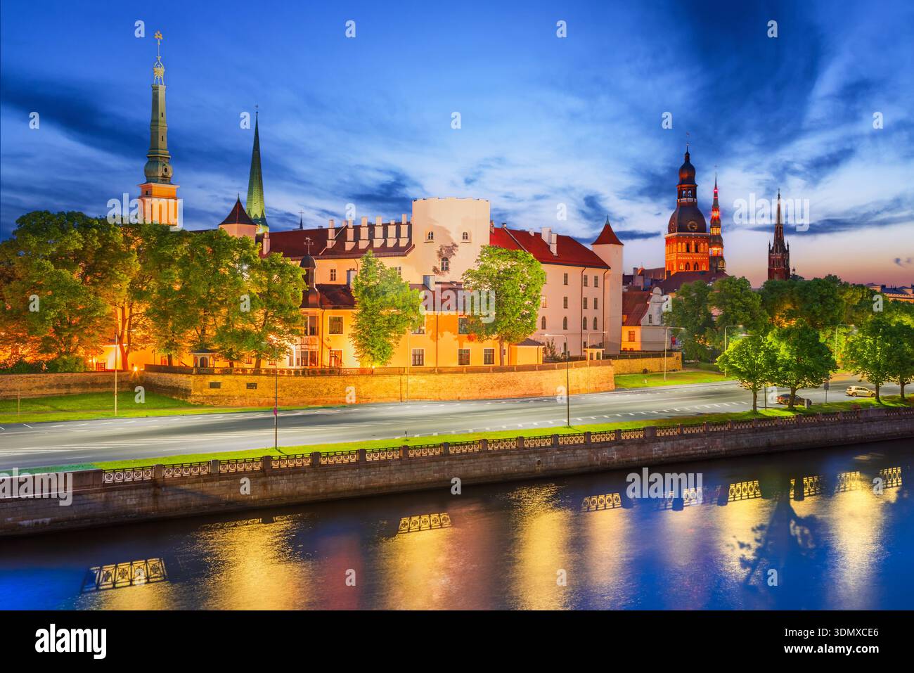 Riga, Lettonia. Scena al crepuscolo della storica città europea lungo il lungofiume Daugava, luci calde che brillano dalla vecchia cittadella e dalle torri della chiesa. Foto Stock