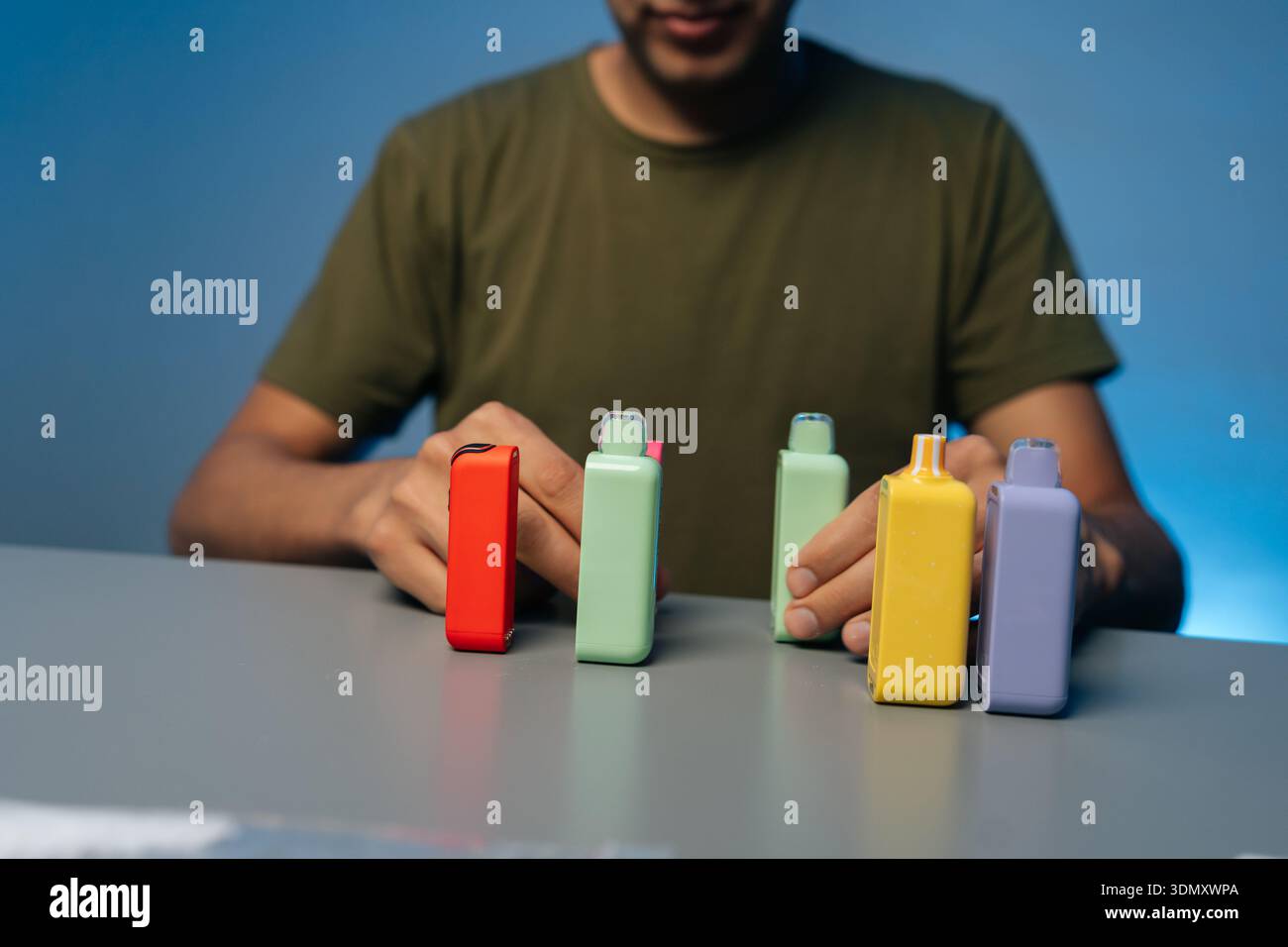 Immagine ritagliata di un uomo irriconoscibile che tiene in mano e presenta vari nuovi sistemi di riscaldamento del tabacco senza bruciature in colori vivaci su un tavolo grigio Foto Stock