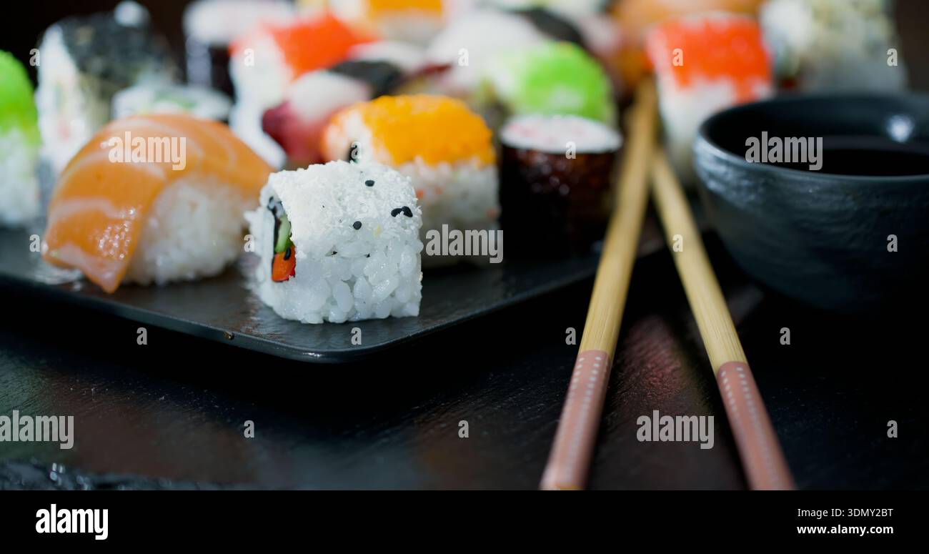 Gli involtini di sushi sono disposti su un piatto nero con bacchette posizionate accanto a loro a un tavolo da pranzo Foto Stock