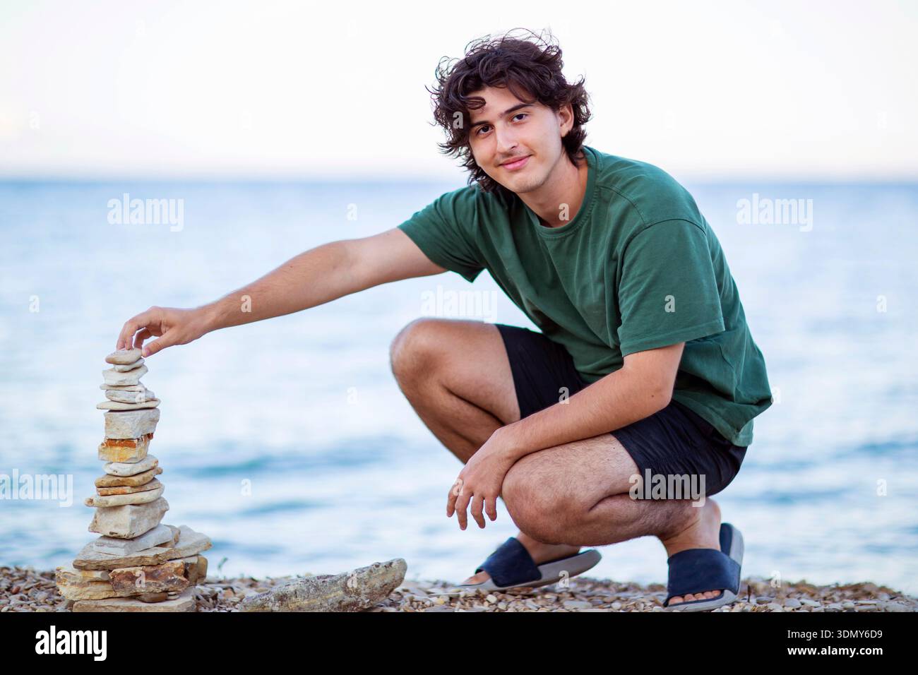 Un giovane uomo che indossa una camicia verde e pantaloncini neri si inginocchia su una spiaggia di ciottoli, impilando con cura le pietre per creare un cairn equilibrato, con il mare sereno Foto Stock