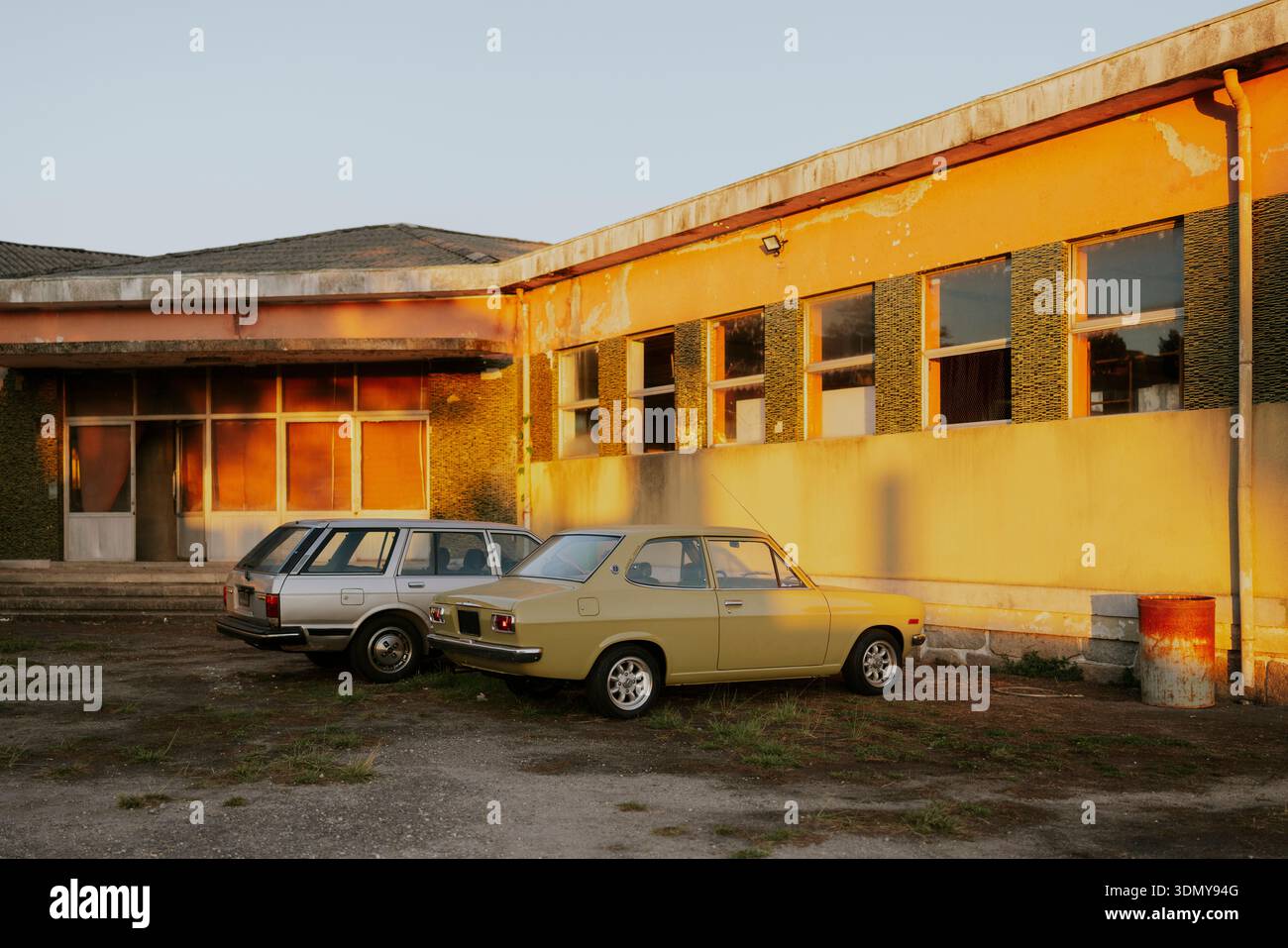 Due auto d'epoca, una gialla e una argento, sono parcheggiate accanto a un edificio abbandonato e intempestivo. Il caldo bagliore del tramonto esalta il nostalgico bancomat Foto Stock