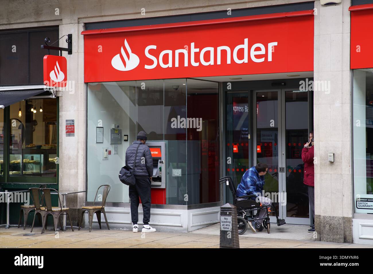 Persone all'ingresso della banca di Santander e bancomat sulla strada della città, incluso l'uomo che ritira contanti e l'utente in sedia a rotelle al telefono. Cambridge, Inghilterra Foto Stock