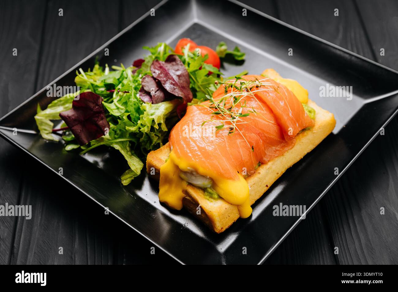 Uova di salmone toast alla benedict con hollandaise e insalata verde su piatto nero, tavolo scuro Foto Stock