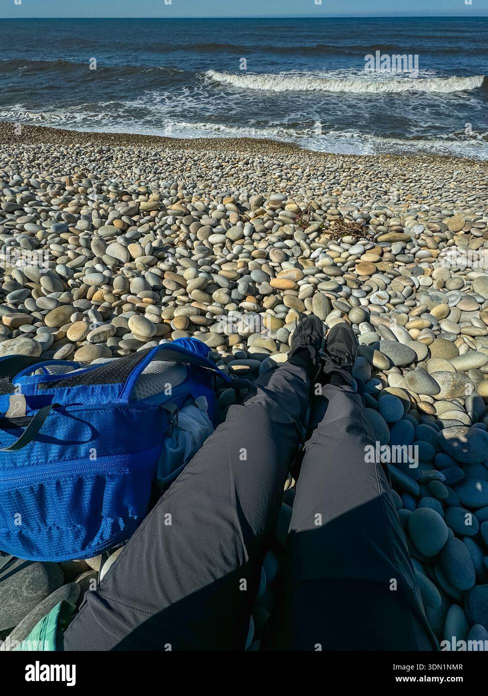 Punto di vista di un viaggiatore solitario che riposa sulla spiaggia di ciottoli con lo zaino e le onde dell'oceano davanti. Pausa durante l'escursione sul cammino di Santiago, viaggio lento Foto Stock