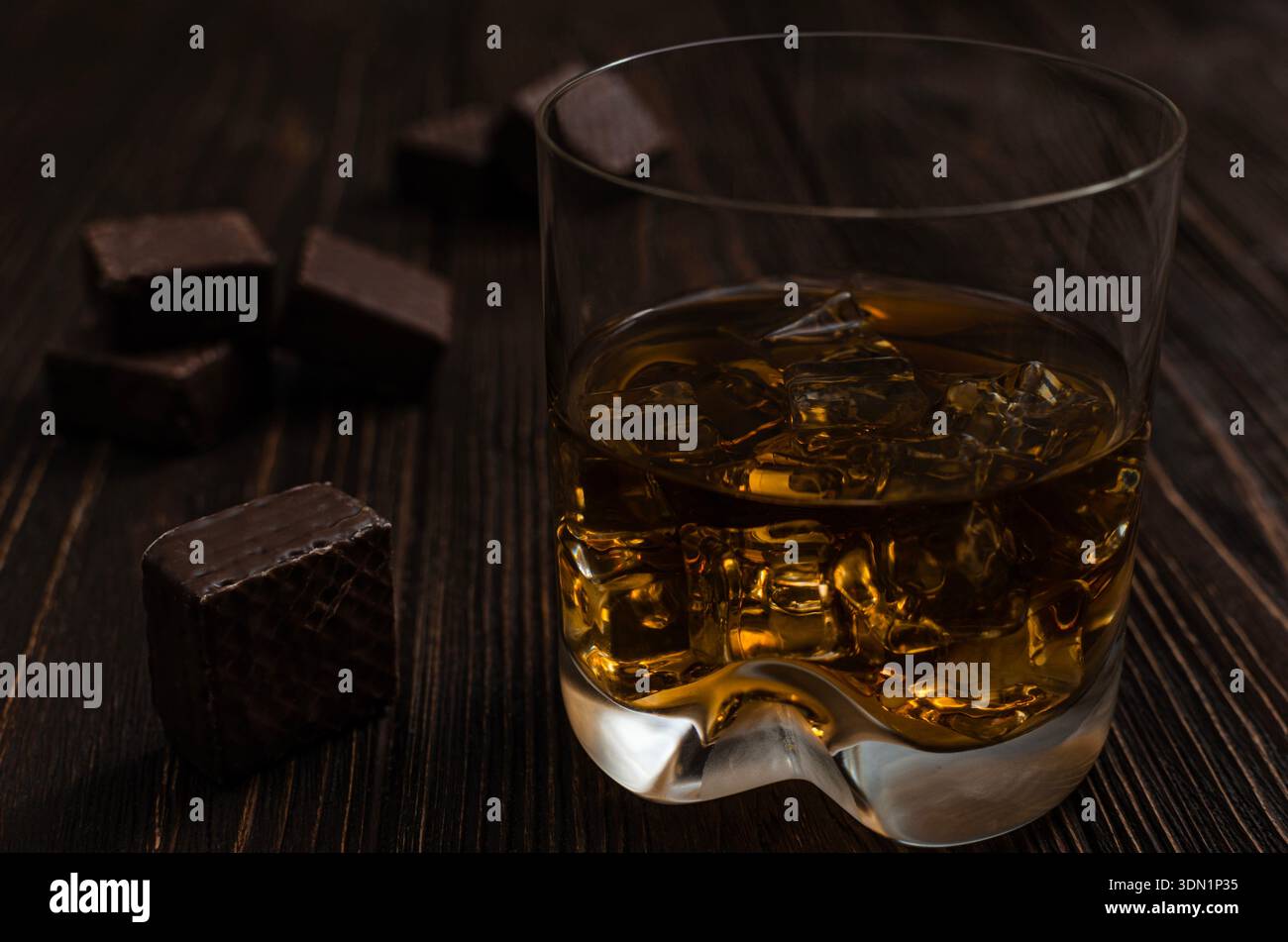 Primo piano di un bicchiere di whisky ambrato con cubetti di ghiaccio, accompagnato da praline di cioccolato fondente su uno sfondo rustico in legno. Foto Stock