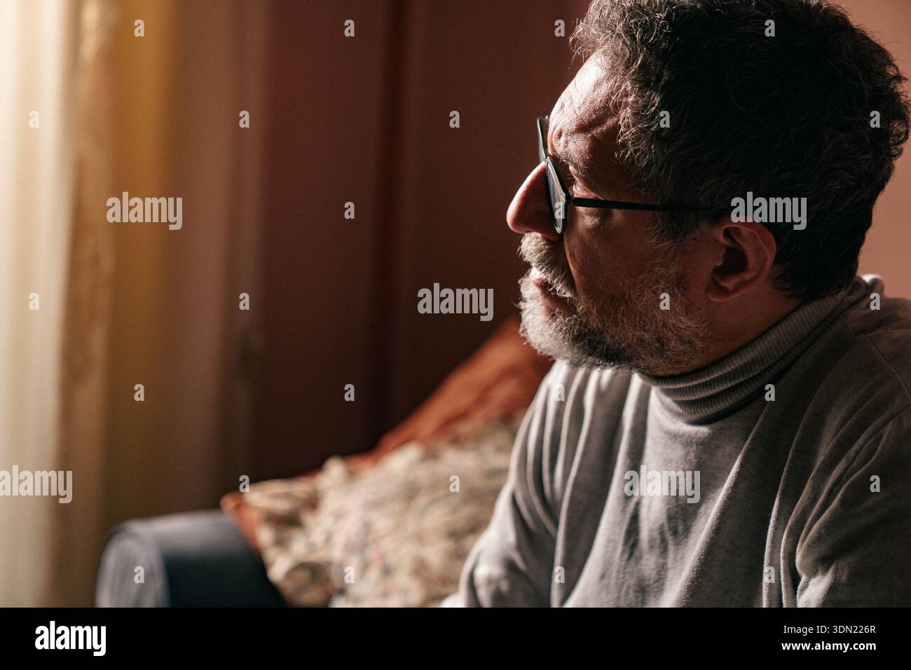 Uomo di mezza età seduto su un divano a casa, riposando la faccia sulla mano mentre pensa. Ambiente interno silenzioso con luce calda, che esprime contemplazione, Foto Stock