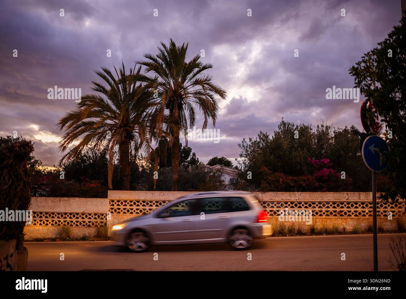 Un'auto argentata catturata con movimento sfocato mentre passa davanti a un muro decorativo in pietra e a due grandi palme contro un vivace cielo nuvoloso al tramonto a Benidorm. Foto Stock