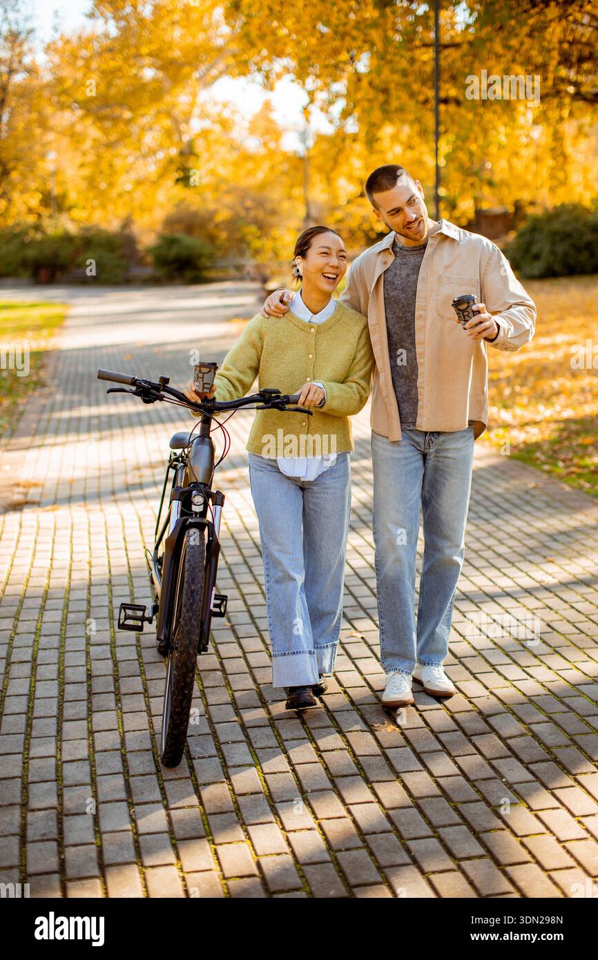 Una donna coreana e un uomo caucasico si godono una allegra passeggiata nel parco. Gli alberi sono adornati con foglie dorate, creando una calda atmosfera autunnale. Foto Stock