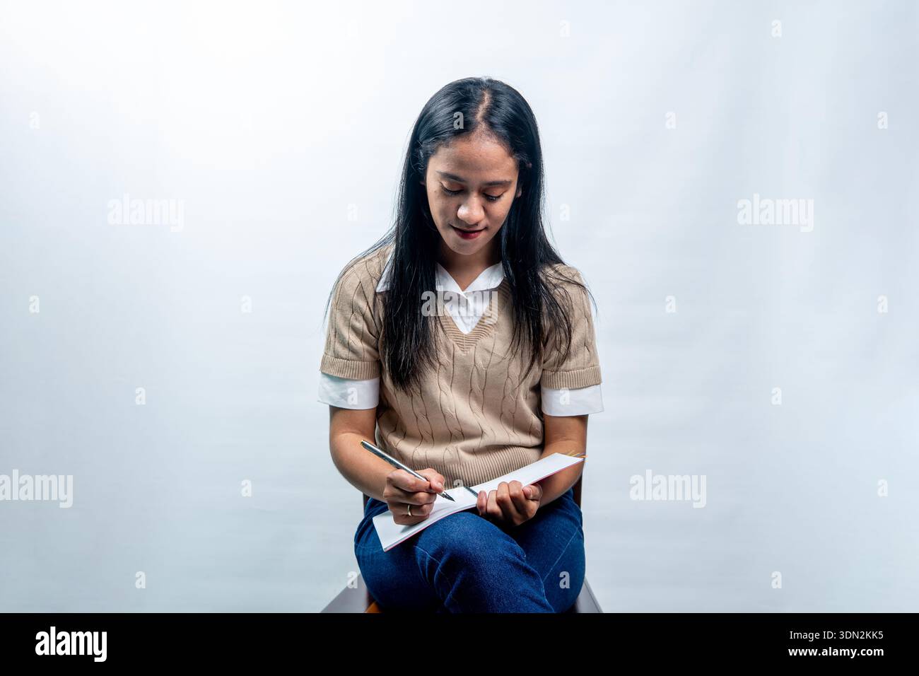 Giovane donna indonesiana calma con un abbigliamento casual e elegante che scrive su un taccuino. Concetto di studio, inserimento nel Journal o pianificazione su uno sfondo bianco pulito. Foto Stock