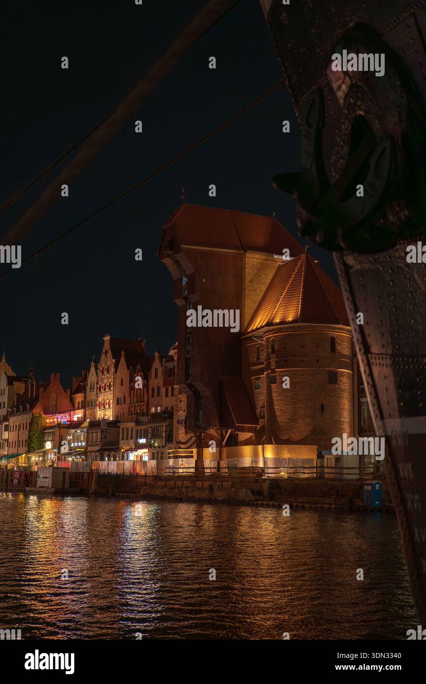 La gru medievale di Danzica sul fiume Motlawa in Polonia è illuminata di notte, con case a schiera a gradini, riflessi increspati e un'ancora ormeggiata Foto Stock