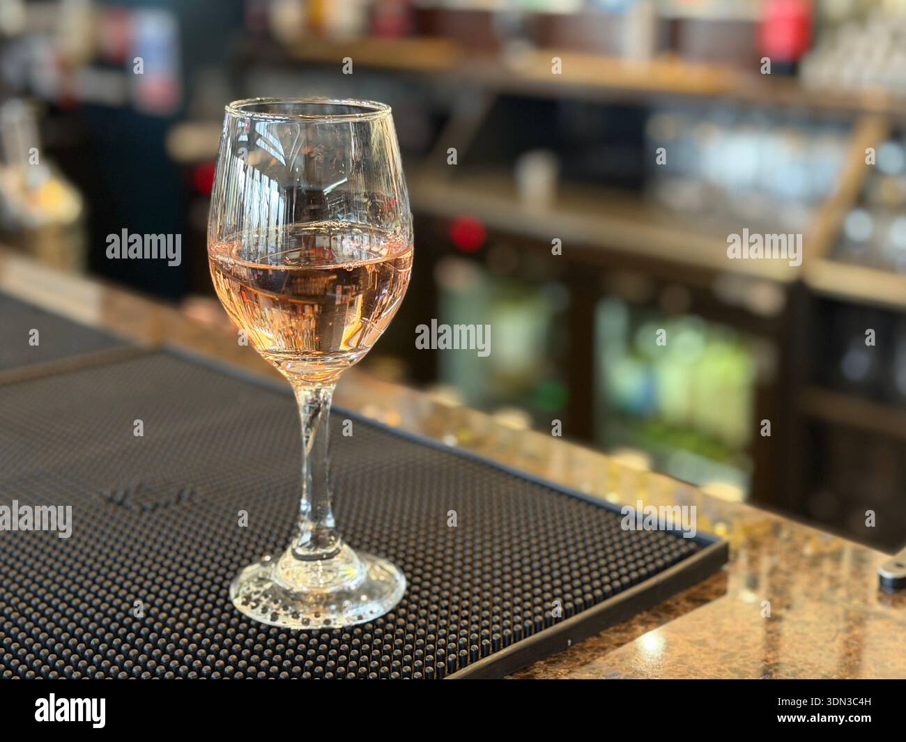 Bicchiere di vino rosato su un bancone bar, con bottiglie e scaffali leggermente sfocati sullo sfondo, in un concetto informale di bevande e ospitalità. Foto Stock