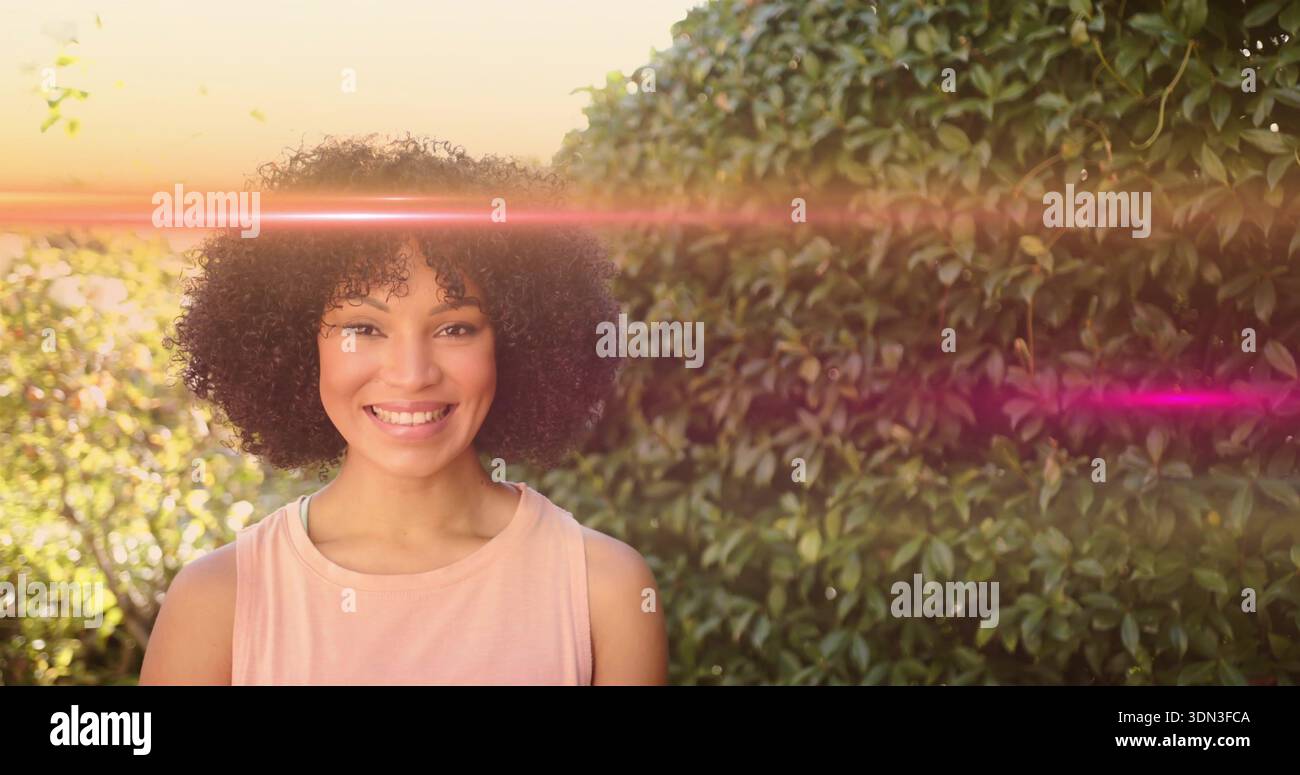 Donna adulta sorridente che indossa una parte superiore rosa chiaro senza maniche in piedi nel giardino sul retro, con luce parassita Foto Stock