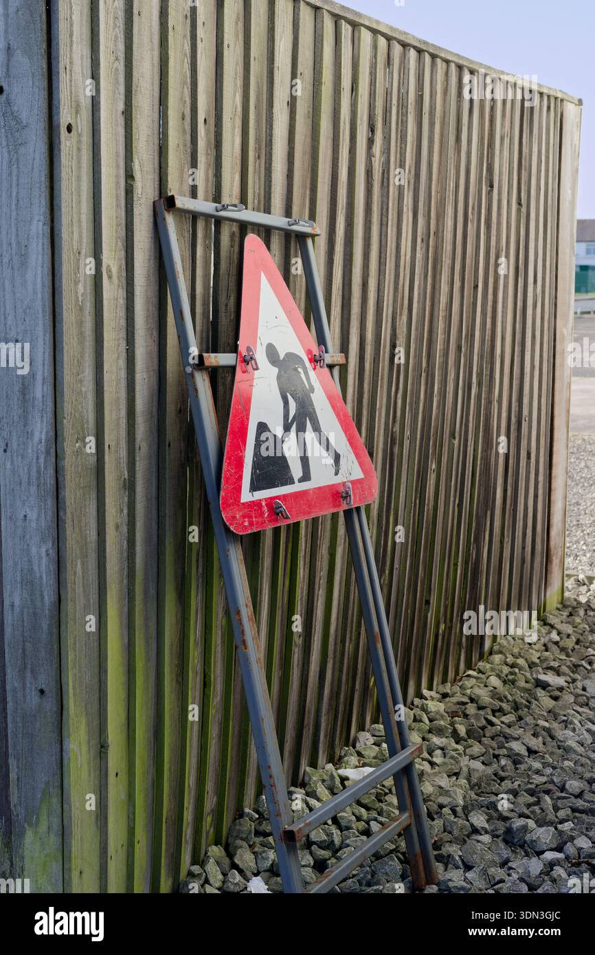 Cartello di lavoro triangolare appoggiato su una recinzione di legno vicino a un sentiero di ghiaia. Il cartello indica gli interventi di manutenzione. Foto Stock
