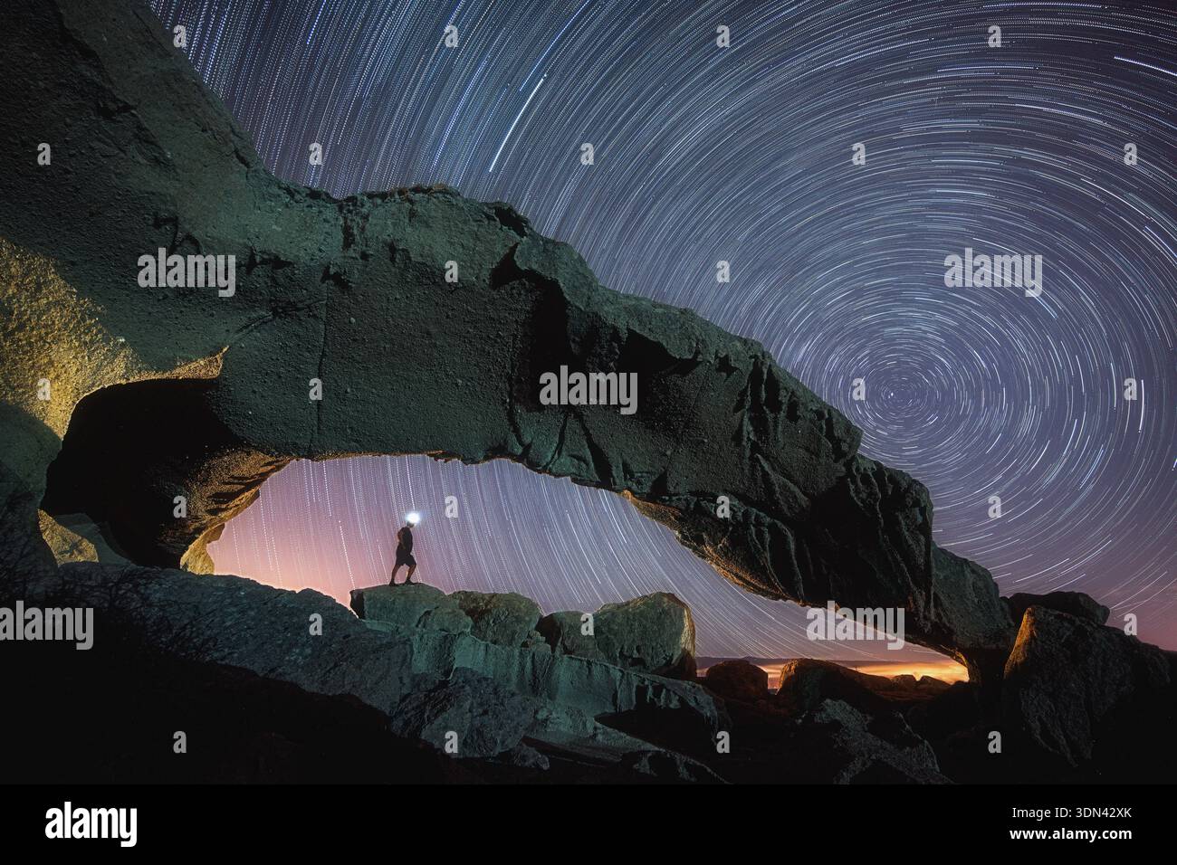 Vista di una figura solitaria che si trova sotto un arco naturale di roccia rocciosa contro un vorticoso cielo stellato, illuminato da un singolo faro, Tajao, CA Foto Stock