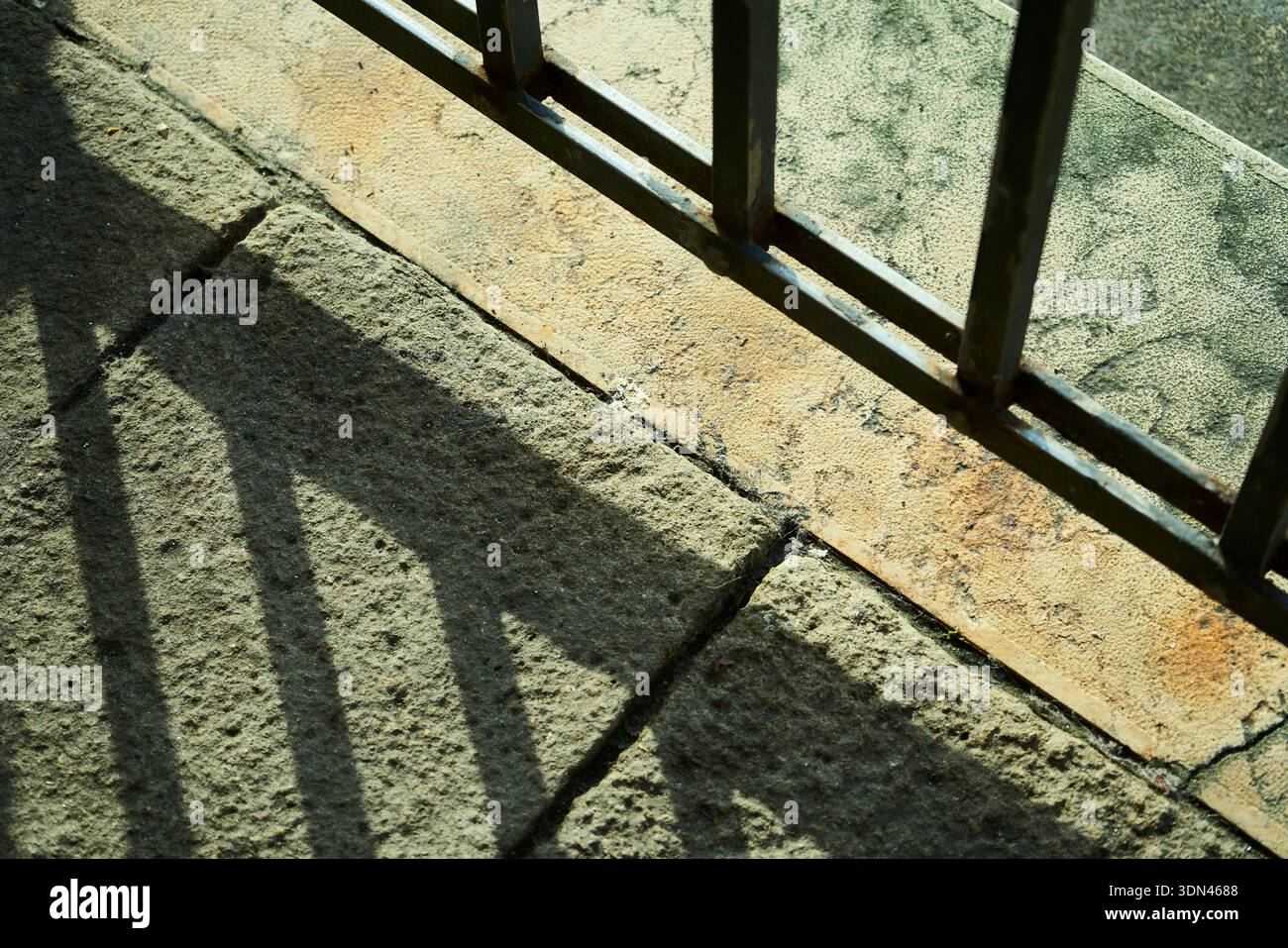 La luce del sole ad alto contrasto crea ombre geometriche da una ringhiera in metallo su un pavimento in pietra antica, creando un motivo astratto minimalista. Foto Stock