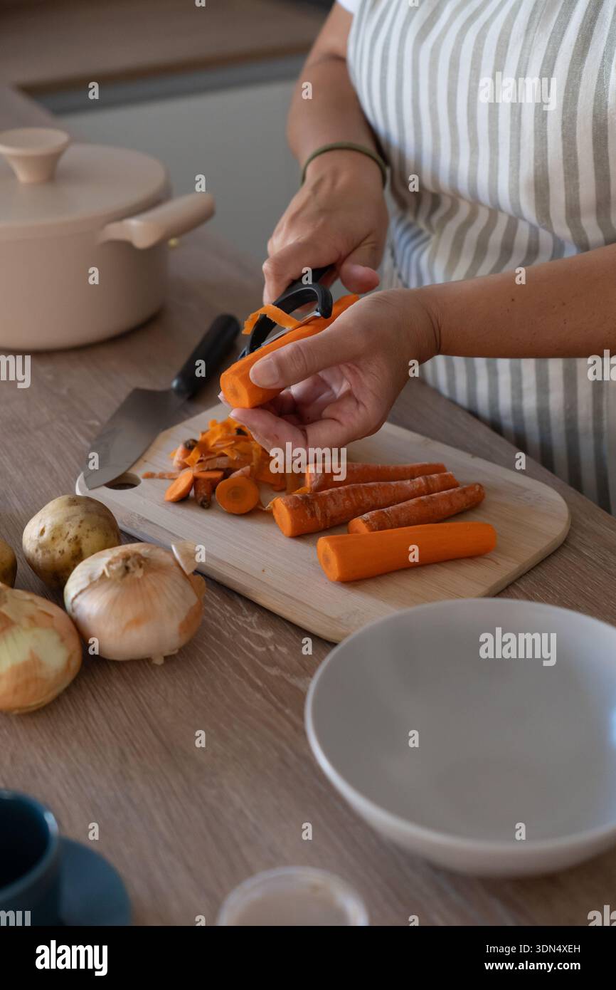 Immagine verticale di mani che sbucciano la carota nella cucina moderna. Cucina casalinga informale, ingredienti freschi e routine quotidiana. Foto Stock