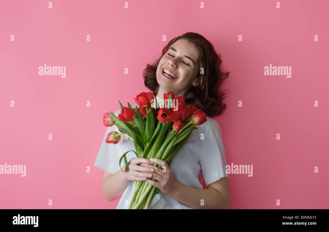 Deliziati con la gioia di questa giovane donna che regge un vivace bouquet di tulipani rossi, adagiato su un vivace sfondo rosa. Il suo sorriso e il suo post rilassato Foto Stock
