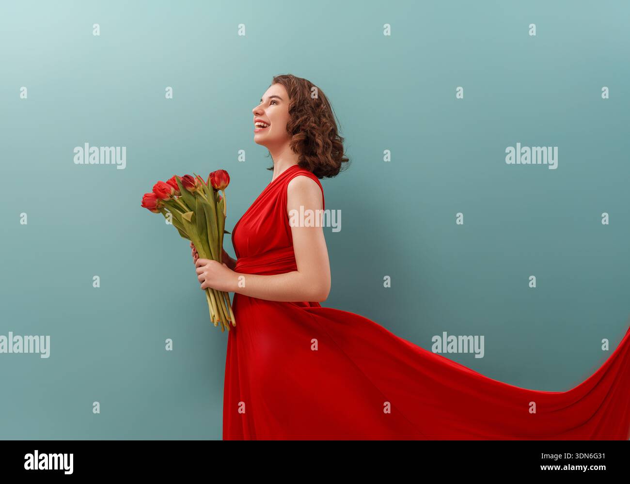 Giovane donna radiosa sorridente con gioia mentre tiene in mano un bouquet di tulipani rossi, vestito con un abito rosso fluente. Lo sfondo minimalista dell'acqua sottolinea Foto Stock