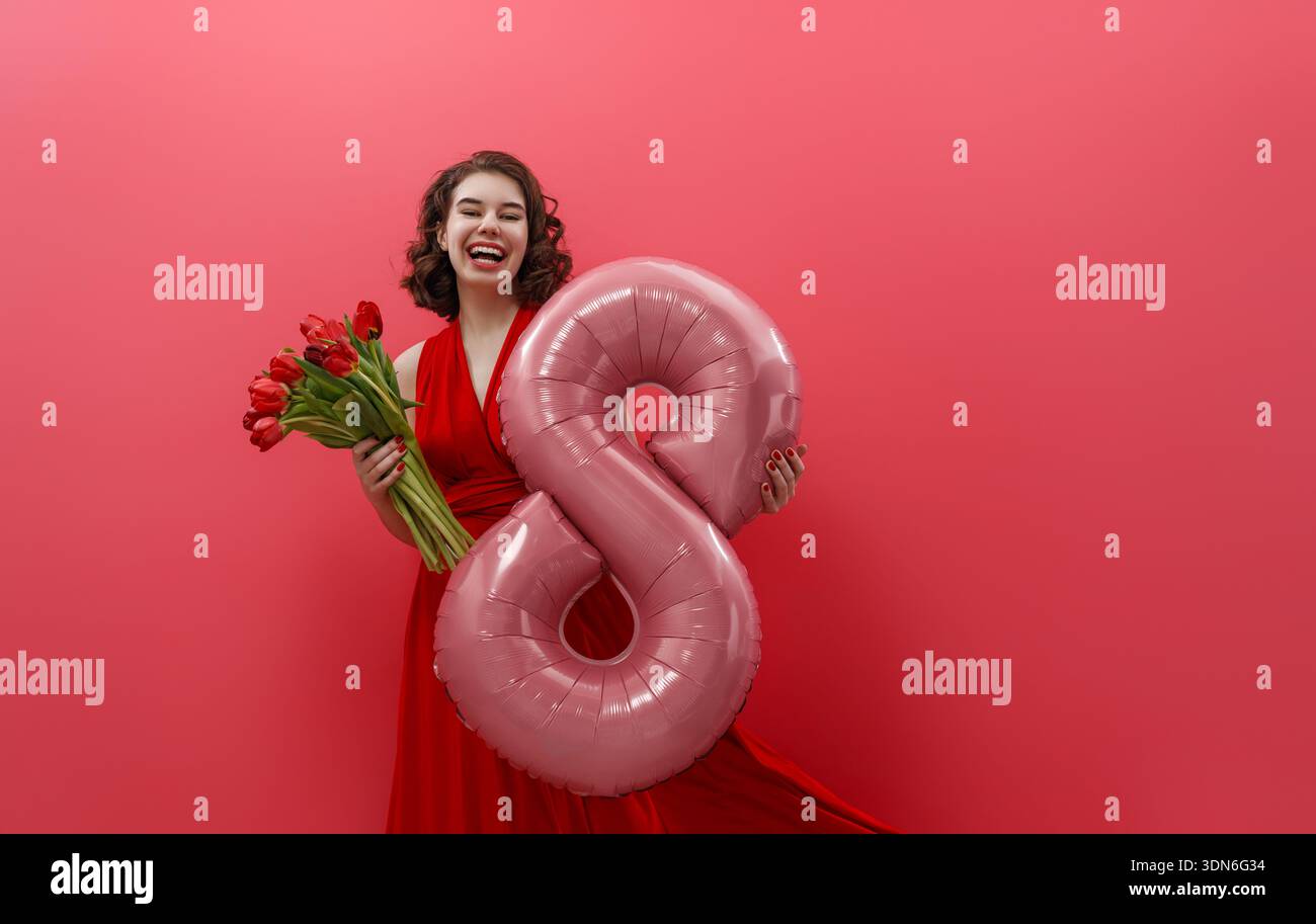 Giovane donna felice in un sorprendente abito rosso che regge con gioia un bouquet di tulipani rossi e un palloncino rosa numero otto, che celebra le donne internazionali Foto Stock