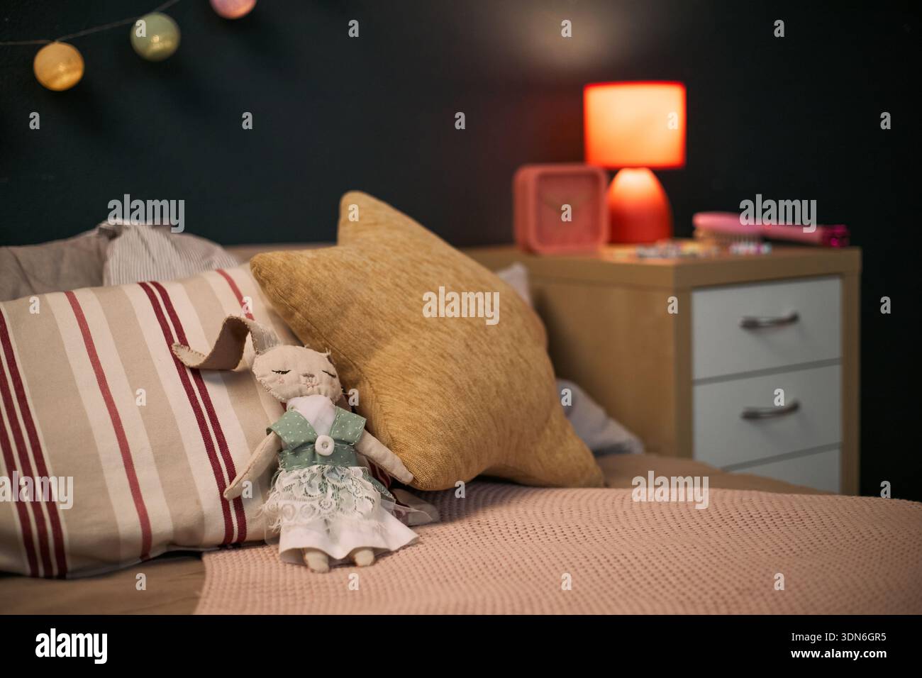 Giocattolo per conigli imbottito che indossa un vestito appoggiato sul letto con cuscini a righe e solidi in un'accogliente camera da letto, comodino con lampada e oggetti decorativi visibili sullo sfondo Foto Stock