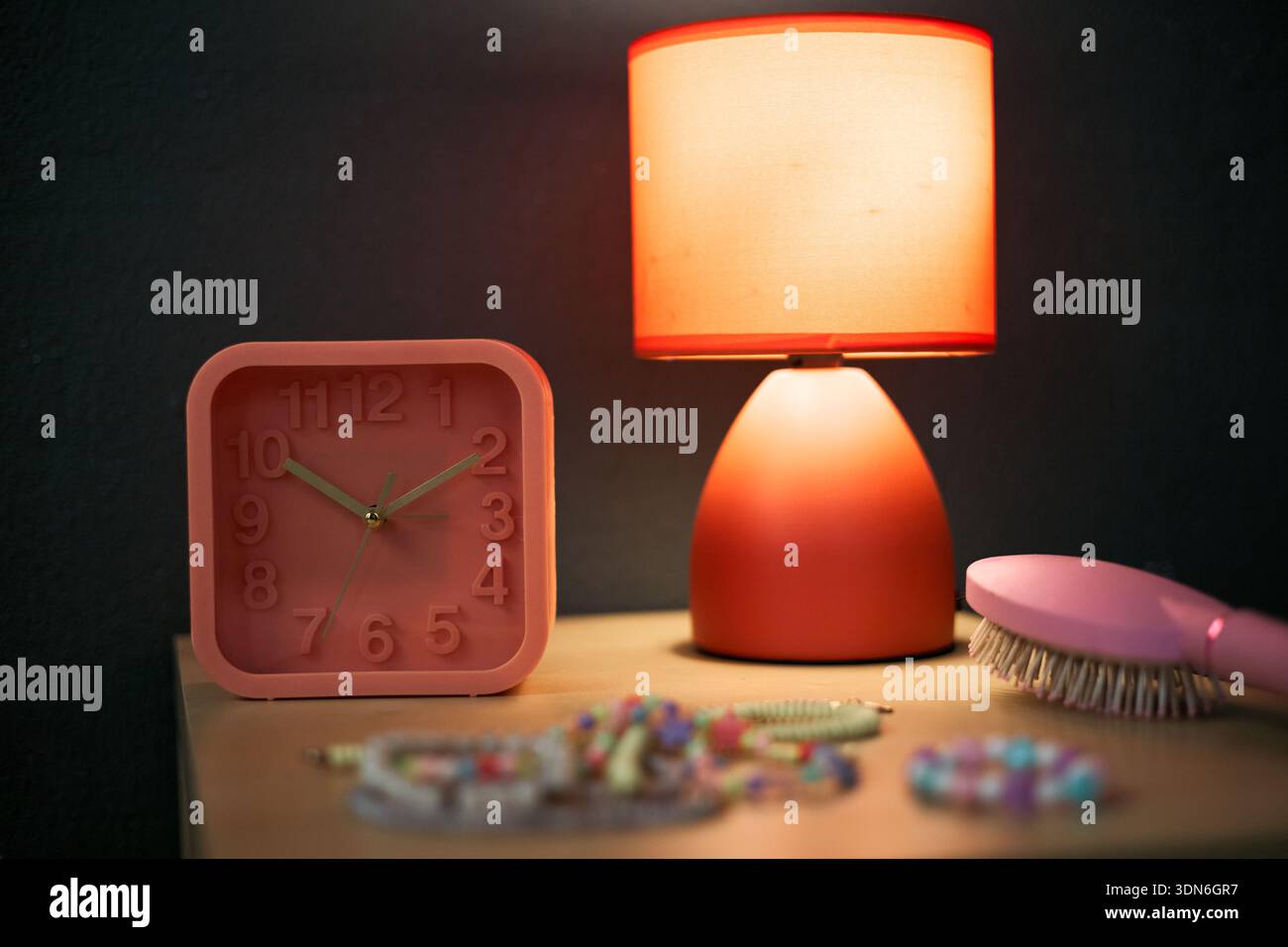 STILL Life mostra sveglia rosa, lampada da tavolo con paralume arancione, spazzola per capelli e gioielli con perline colorate disposte su una superficie in legno, oggetti illuminati da una luce calda Foto Stock