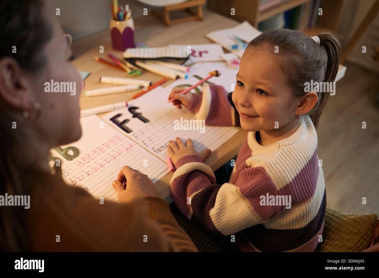 Bambina caucasica sorridente mentre si pratica a scrivere lettere con matita colorata, seduta alla scrivania con donna adulta che assiste durante l'attività di apprendimento, materiali educativi visibili Foto Stock