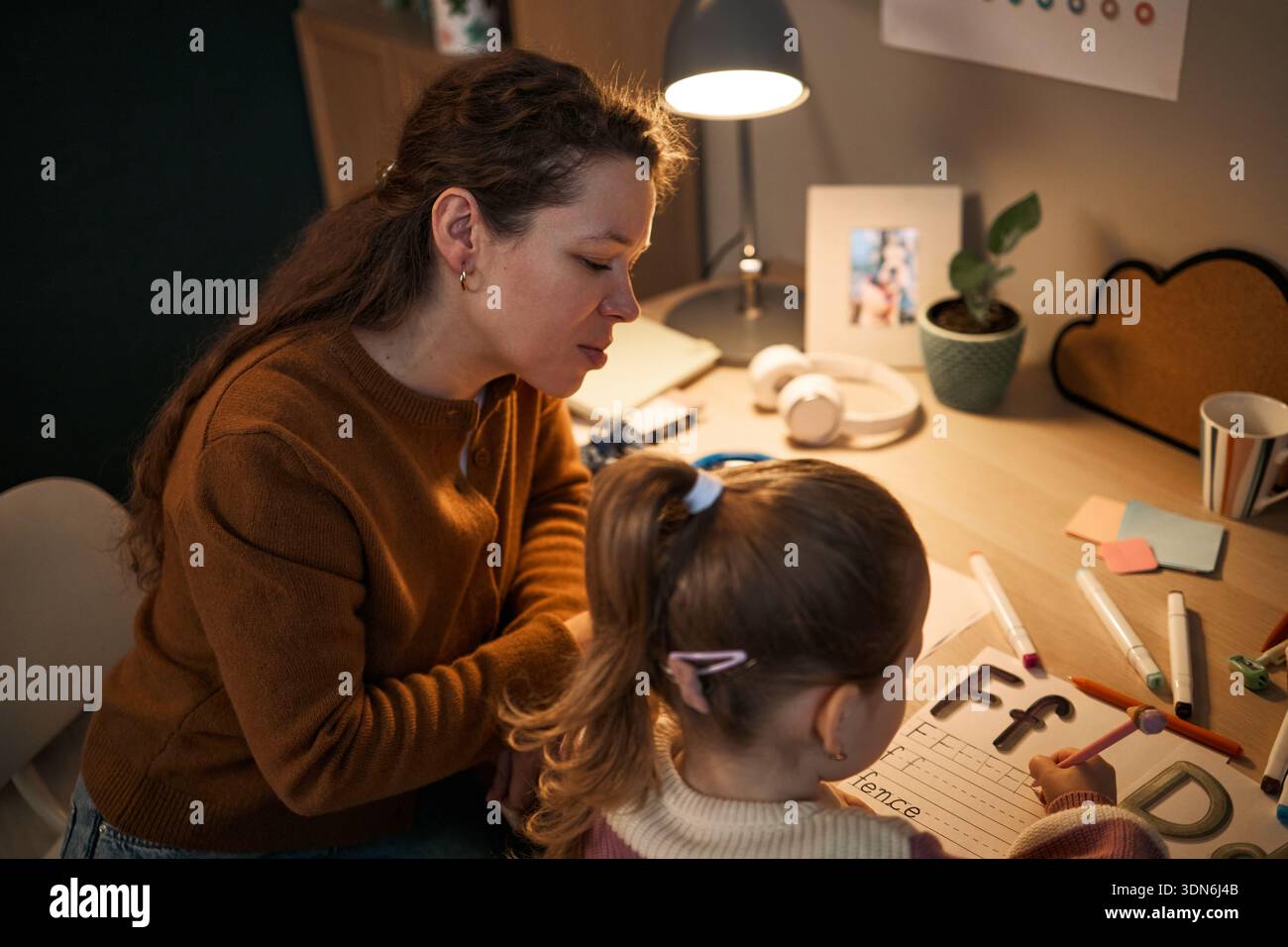 Donna adulta caucasica che aiuta un bambino caucasico con pratica di scrittura a mano alla scrivania, entrambe focalizzate sull'attività di apprendimento, materiali didattici e cancelleria sparsi nello spazio di lavoro Foto Stock