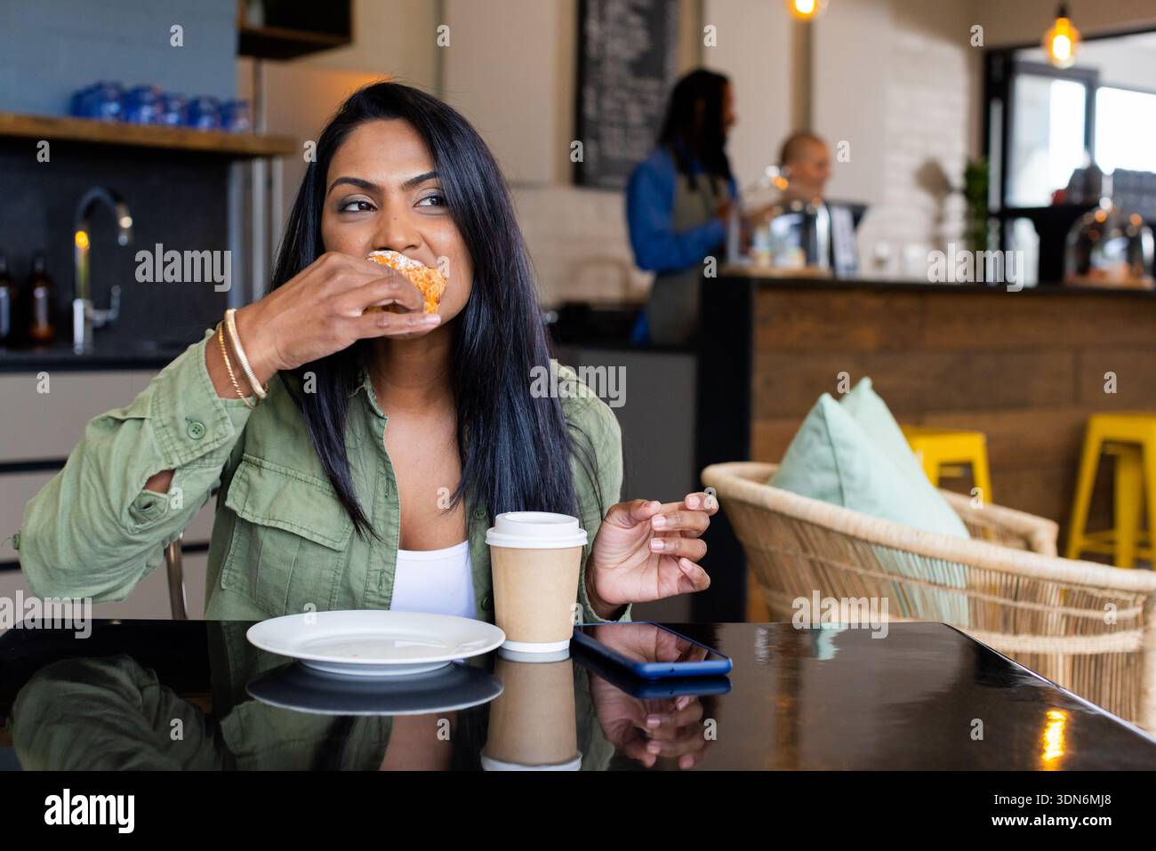 Donna asiatica indiana che morde pasticceria e tiene una tazza di caffè al tavolino nero lucido con smartphone Foto Stock