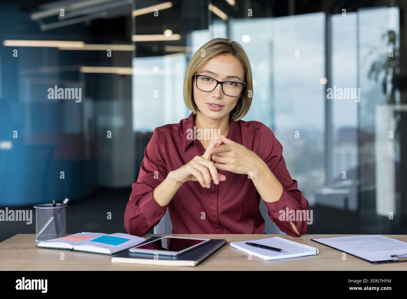 Donna d'affari sicura che indossa occhiali da vista e una camicia marrone seduta alla scrivania, gestando con le mani durante una riunione virtuale in un moderno ufficio open space Foto Stock
