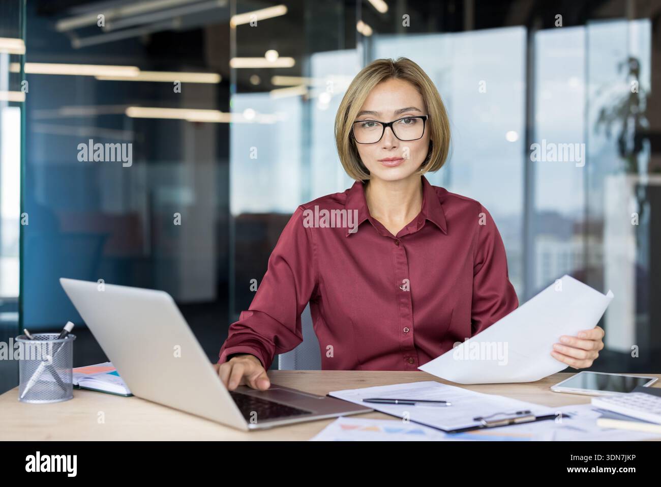 Donna d'affari sicura che indossa occhiali da vista e una camicia marrone, guarda la fotocamera mentre lavora su un laptop e tiene in mano documenti, concentrandosi sulla gestione e sul successo aziendale nel suo ambiente di lavoro moderno Foto Stock