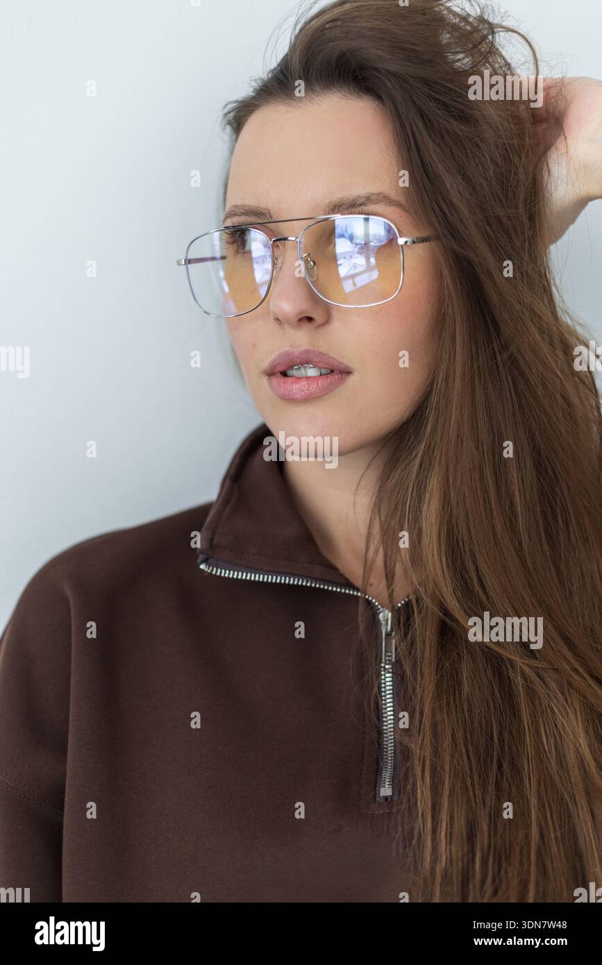 Ritratto di una donna che indossa una felpa marrone alla moda con cappuccio e occhiali in argento. Look rilassato e autentico con illuminazione soffusa e colori neutri. Ideale per blog po Foto Stock