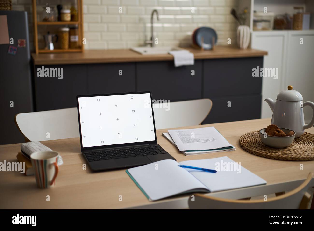 Moderno tavolo da cucina con laptop aperto con schermo vuoto, notebook e penna sparsi, teiera e tazza su tappeto intrecciato, suggerendo un ambiente di lavoro remoto o di studio Foto Stock
