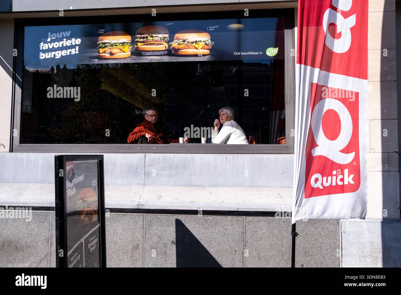 Due clienti anziani siedono a un tavolo all'interno di un fast food dietro uno striscione alla finestra che legge hamburger favoriti Jouw e Veggie al Koningin Foto Stock