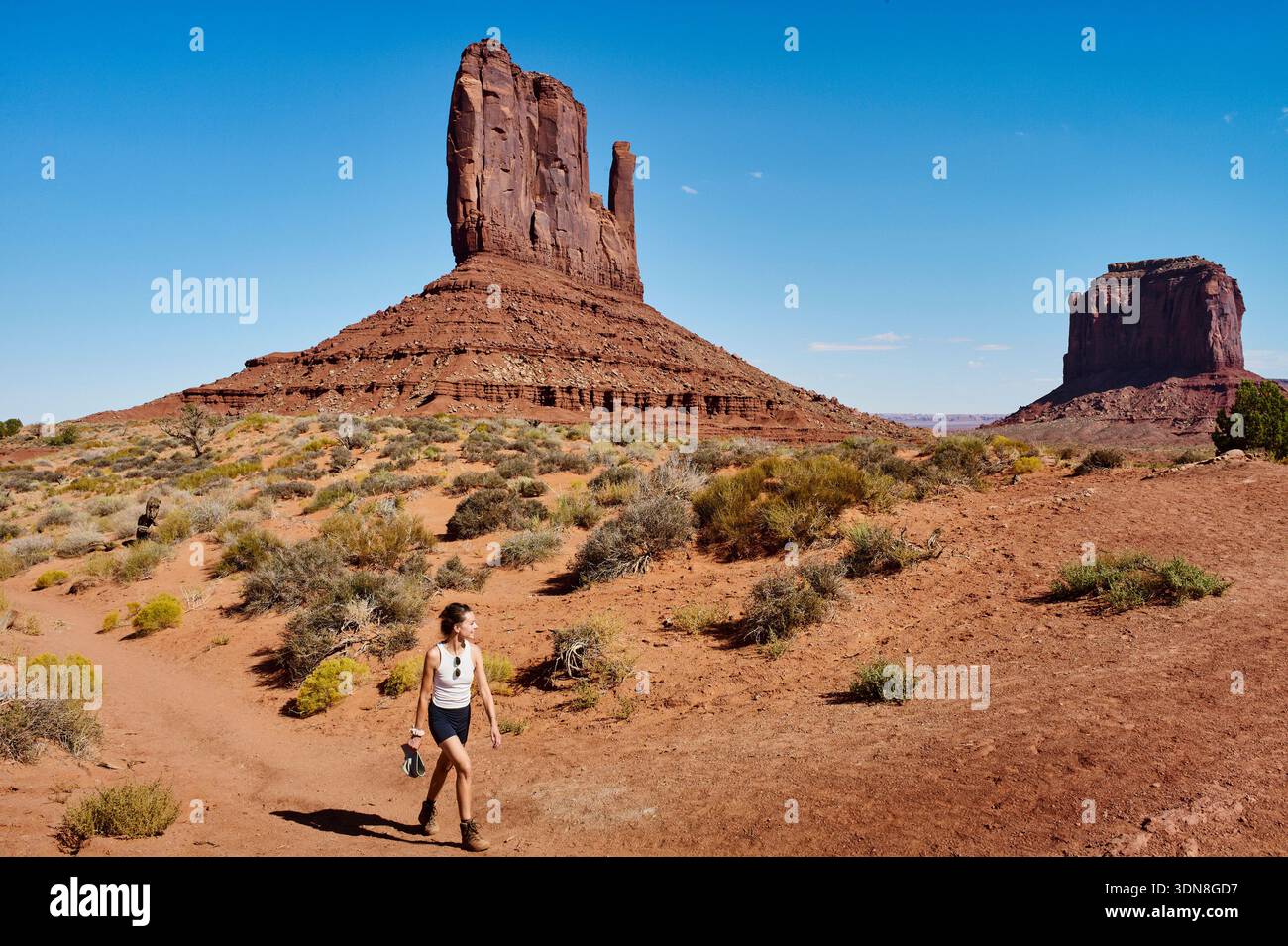 Una donna cammina attraverso un paesaggio desertico con enormi formazioni rocciose sotto un cielo blu brillante. Monument Valley, Arizona, Stati Uniti Foto Stock