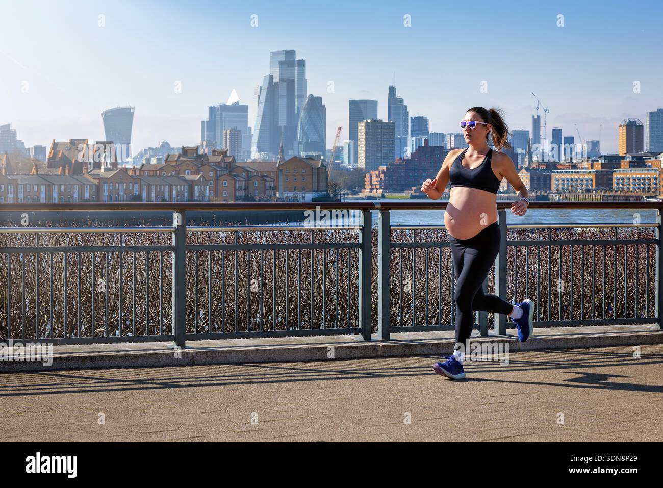 Una donna incinta in abbigliamento sportivo che corre per l'esercizio fisico di fronte a uno skyline urbano durante una giornata di sole Foto Stock