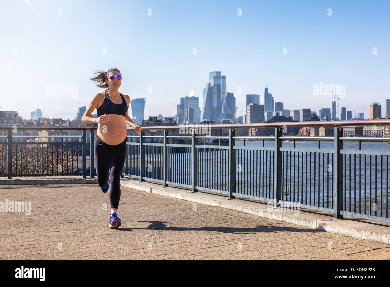 Donna atletica incinta in abbigliamento sportivo che corre per l'esercizio fisico di fronte a uno skyline urbano durante una giornata di sole Foto Stock