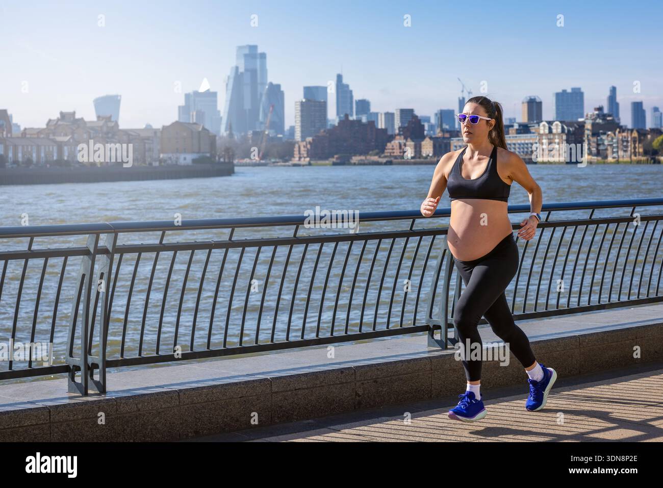 Una donna incinta in abbigliamento sportivo che corre per l'esercizio fisico di fronte a uno skyline urbano durante una giornata di sole Foto Stock