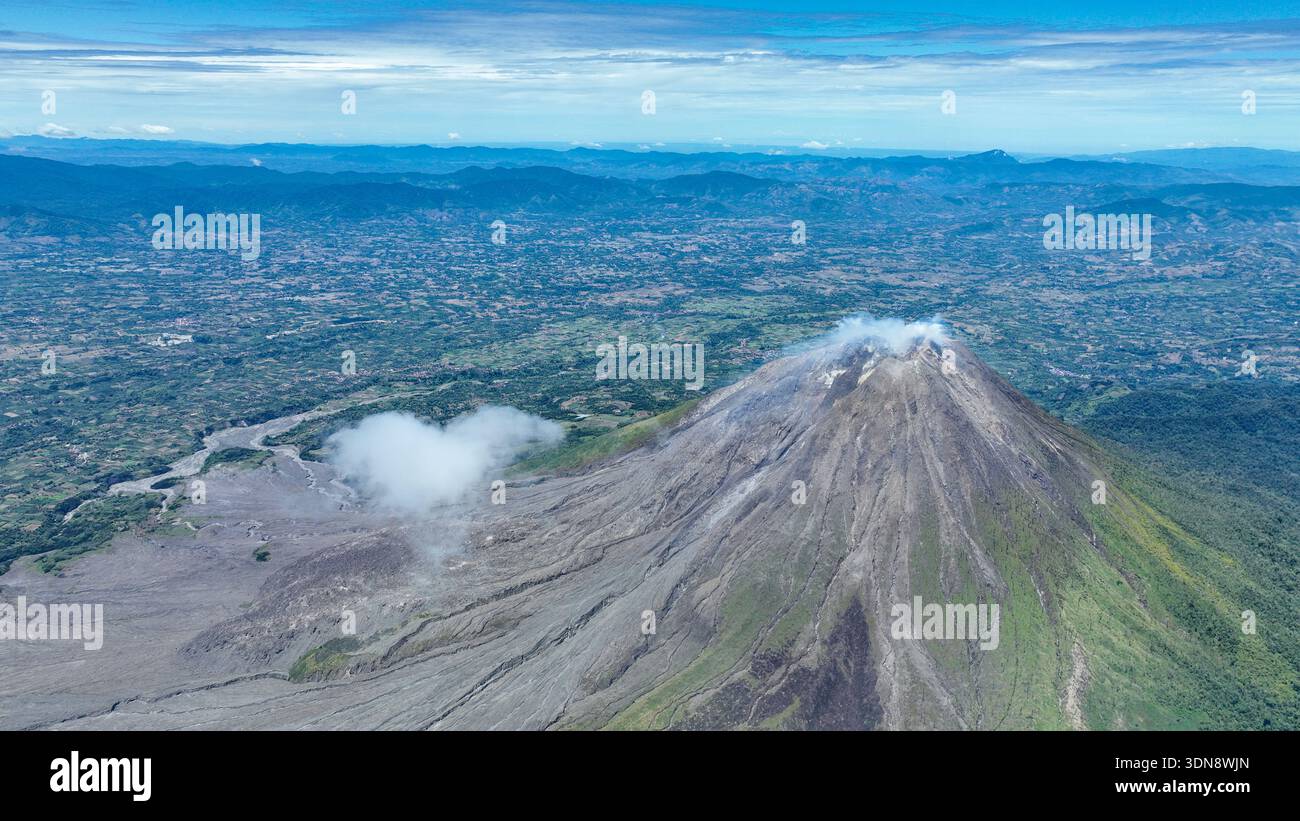 Da questa altezza, Sinabung sembra una gigantesca onda di pietra congelata durante un incidente, fumo che si arriccia dal suo cratere verso le fattorie di patchwork Foto Stock