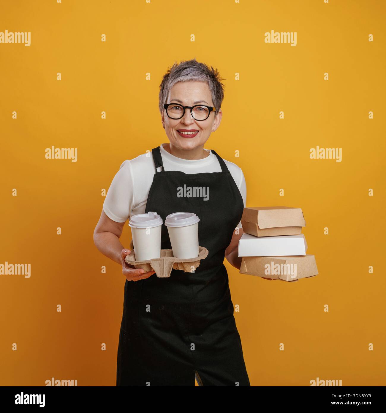 Una donna allegra con capelli e bicchieri grigi corti regge un vassoio con tazze da caffè e scatole da asporto su uno sfondo giallo brillante, che rappresenta Foto Stock