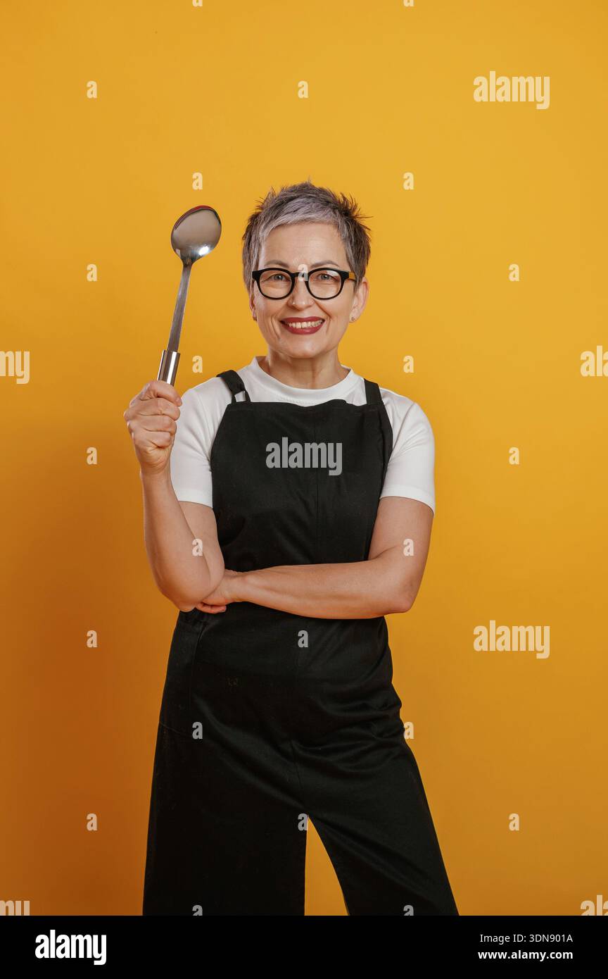 Una donna allegra che cuoca con i capelli corti grigi tiene con sicurezza un cucchiaio da cucina mentre indossa un grembiule nero su un vivace sfondo giallo Foto Stock