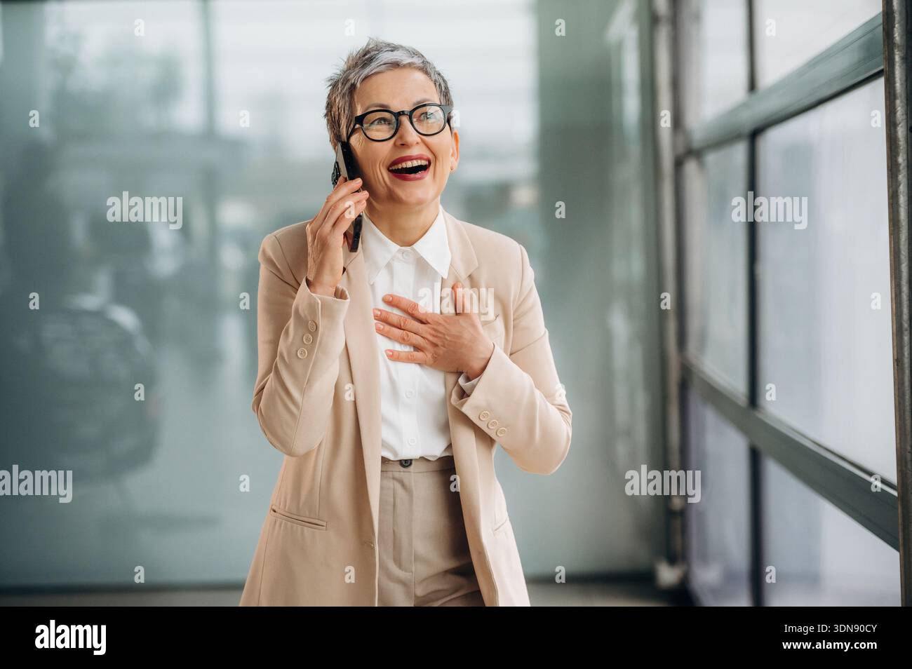 Una donna anziana vestita con un abito alla moda ride gioiosamente mentre parla sul suo smartphone, circondata da un luminoso spazio ufficio con un grande vento Foto Stock