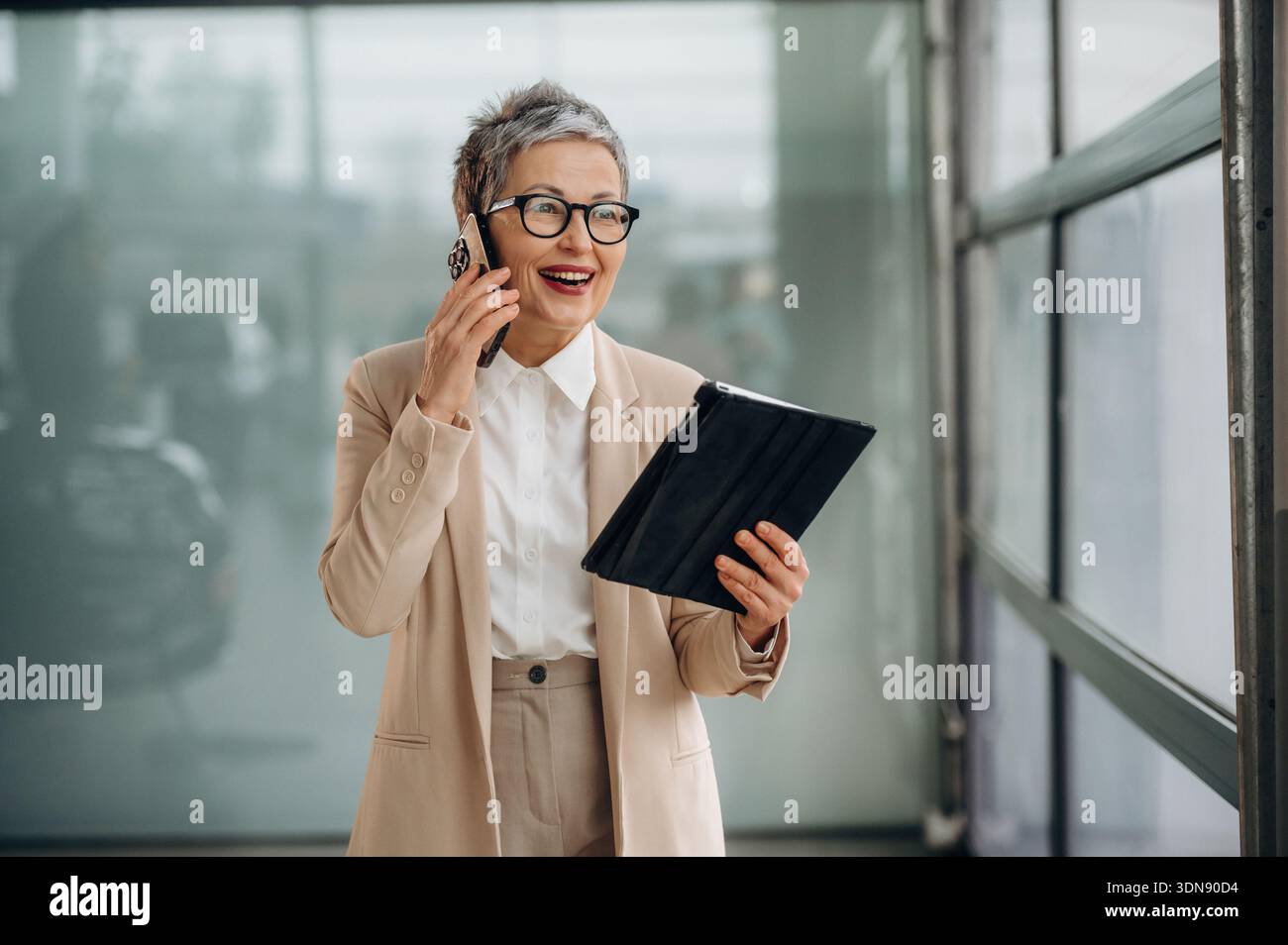 Una donna matura con i capelli grigi corti sorride mentre è impegnata in una conversazione telefonica e tiene in mano un tablet in un moderno ufficio con grandi dimensioni Foto Stock