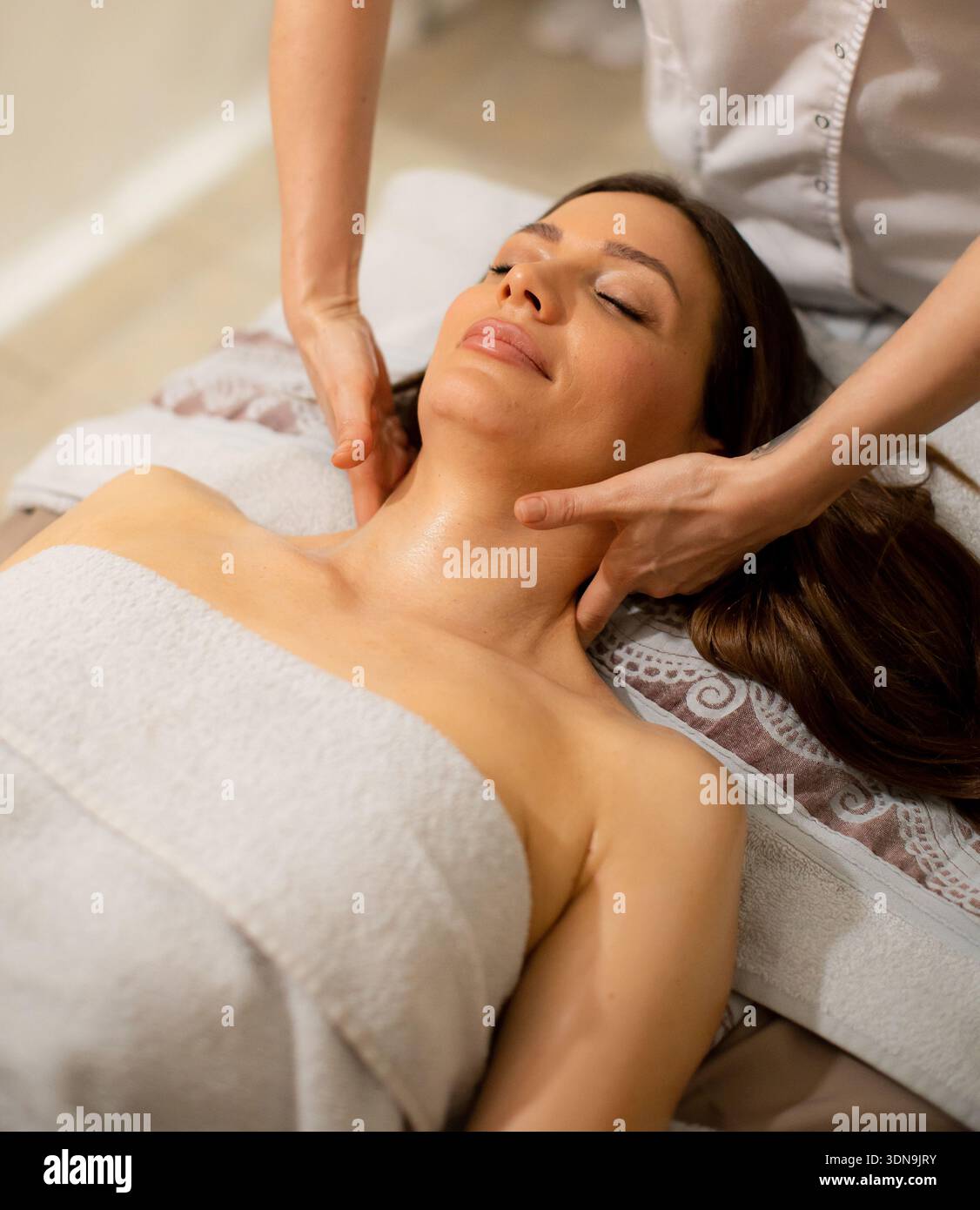 Una donna è sdraiata su un tavolo di trattamento mentre un terapeuta esegue un massaggio al collo. L'ambiente e' una spa con luci soffuse e decorazioni rilassanti. È af Foto Stock