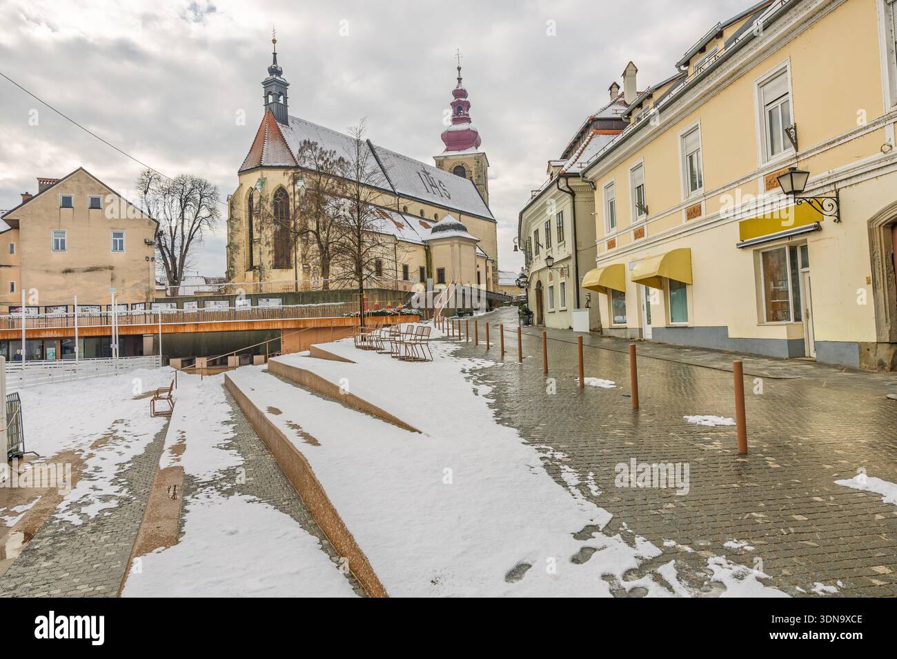 PTUJ, SLOVENIA, 01.04.2026: Una strada urbana innevata presenta una chiesa storica, edifici color giallo pastello e una passeggiata acciottolata. Sedie vuote e guerra Foto Stock