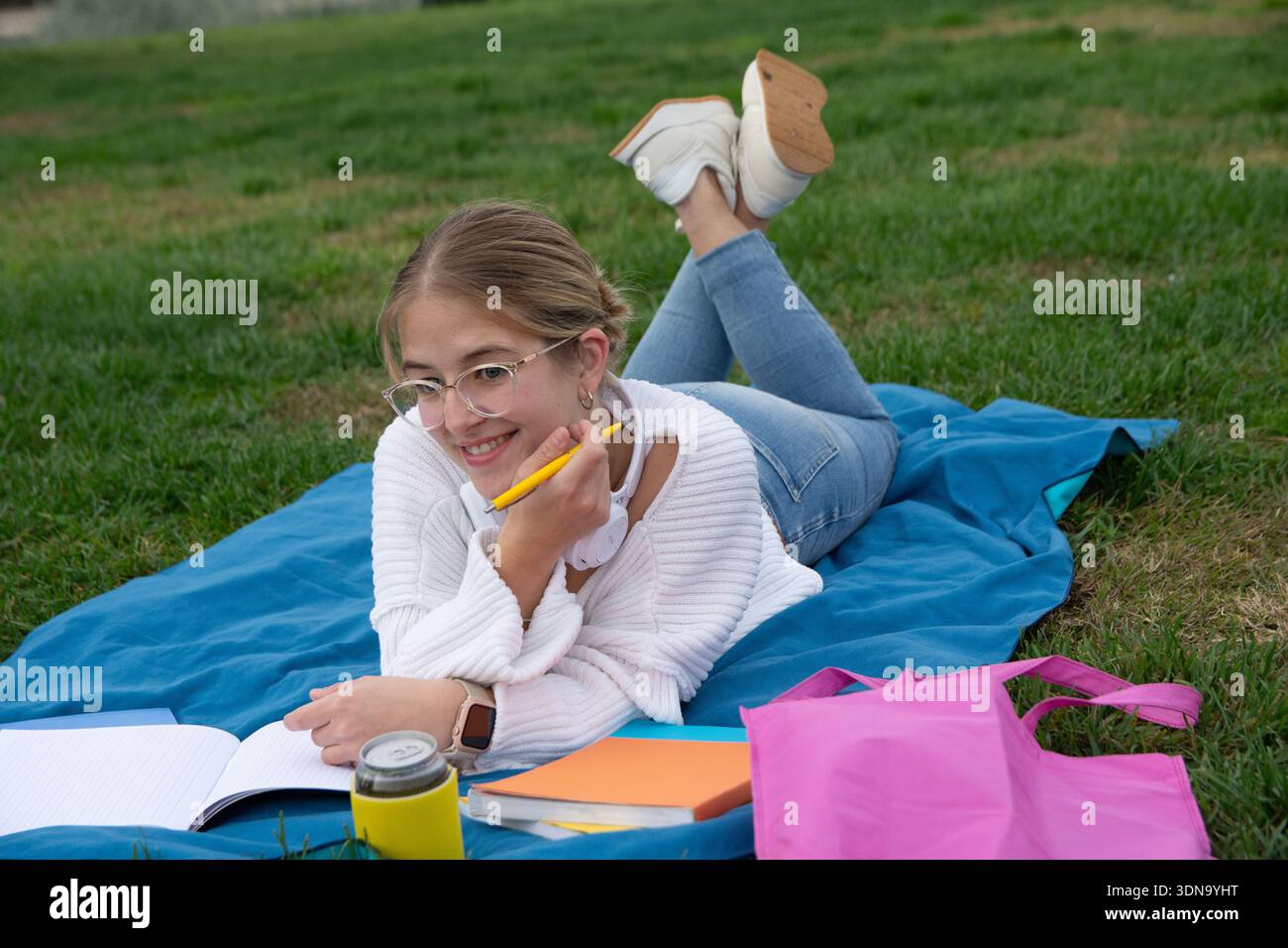 Giovane studentessa sdraiata su una coperta sull'erba del campus universitario, sorridente e concentrata mentre scrive su un taccuino, godendo di una formazione all'aperto Foto Stock