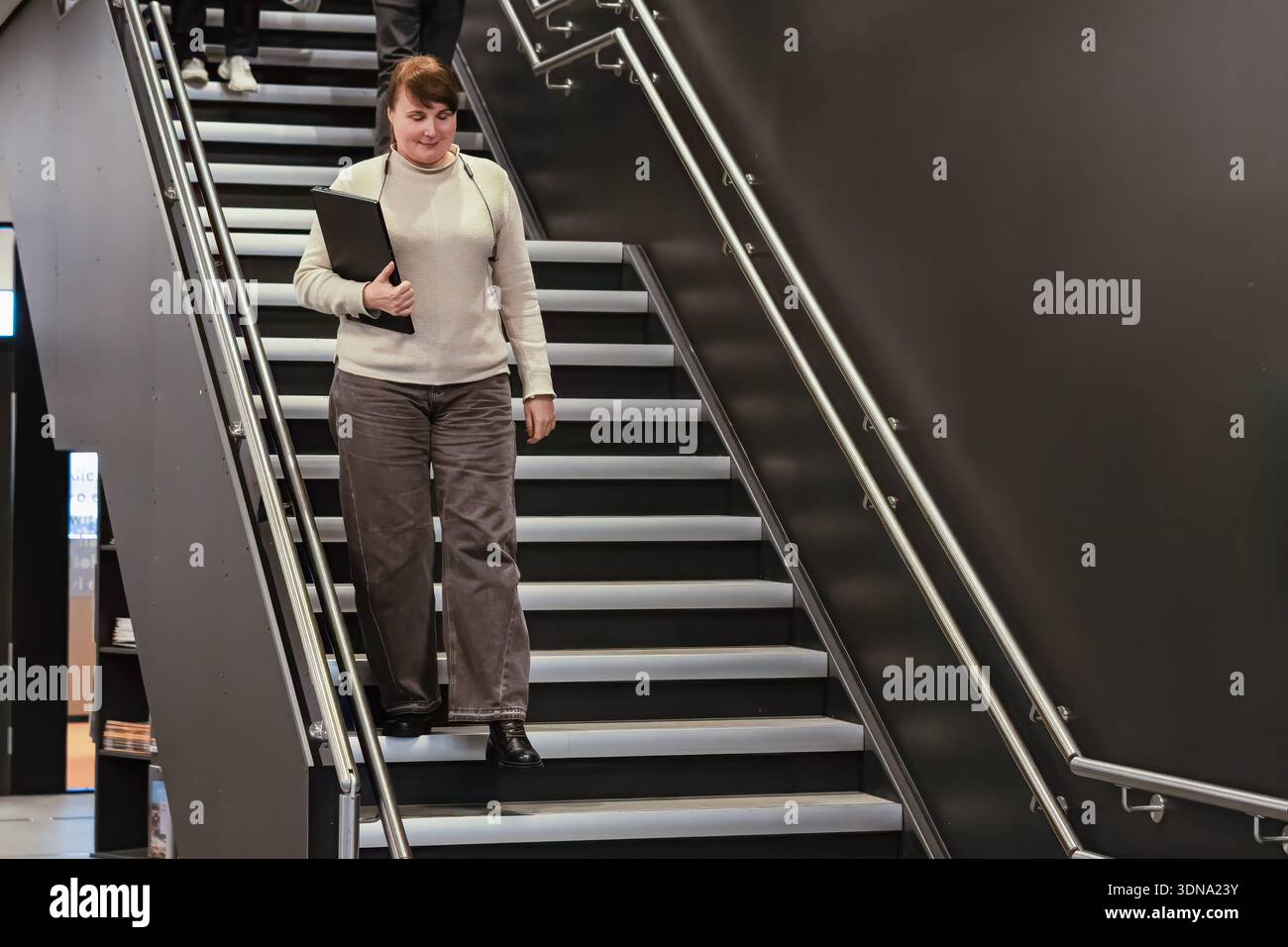 Una donna professionista sta camminando lungo un'elegante scala, tenendo in mano una cartella, circondata da un'architettura moderna e da un'atmosfera elegante, che riflette un dinamismo Foto Stock