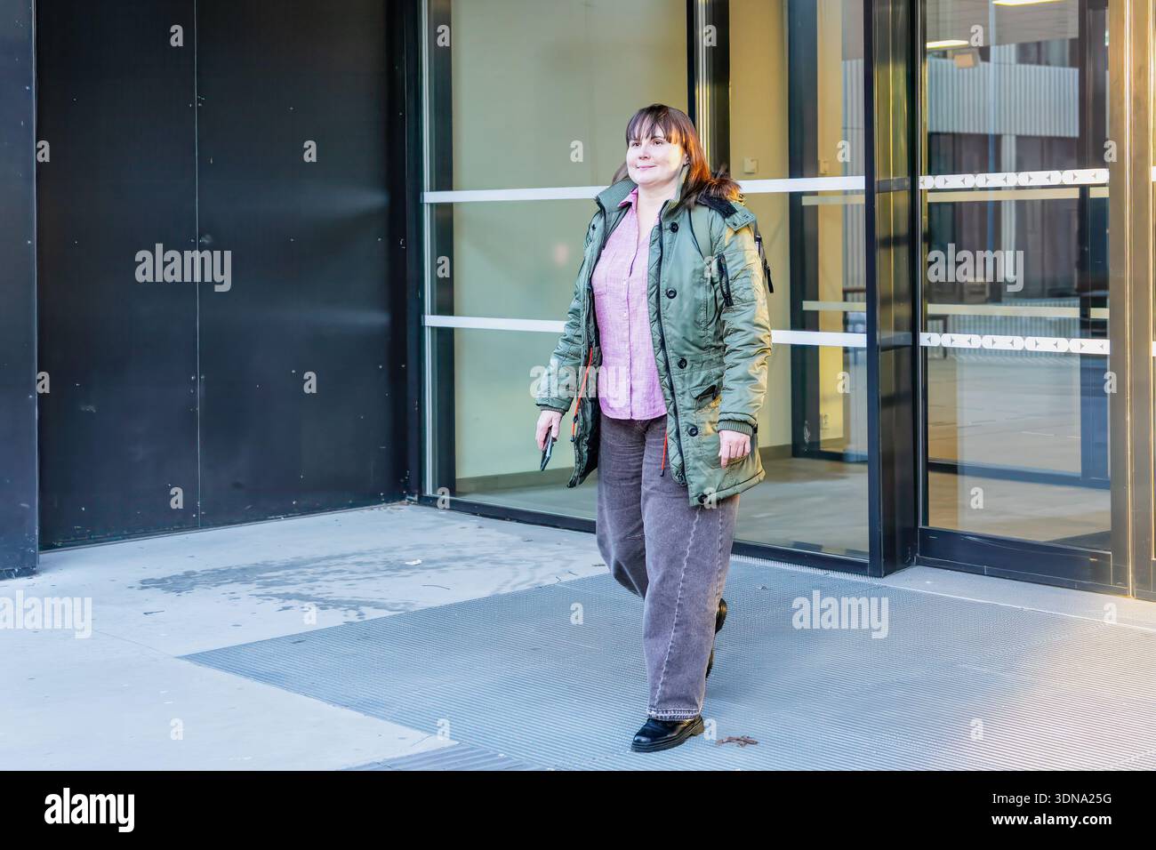 Una donna con giacca verde e pantaloni grigi, cammina fuori da una struttura contemporanea, incarnando lo stile urbano e la fiducia in un ambiente vivace Foto Stock