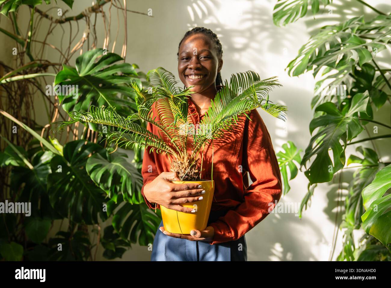 Una donna afroamericana sorridente tiene Cycas in vaso, circondata da lussureggianti giardini tropicali. Foto Stock