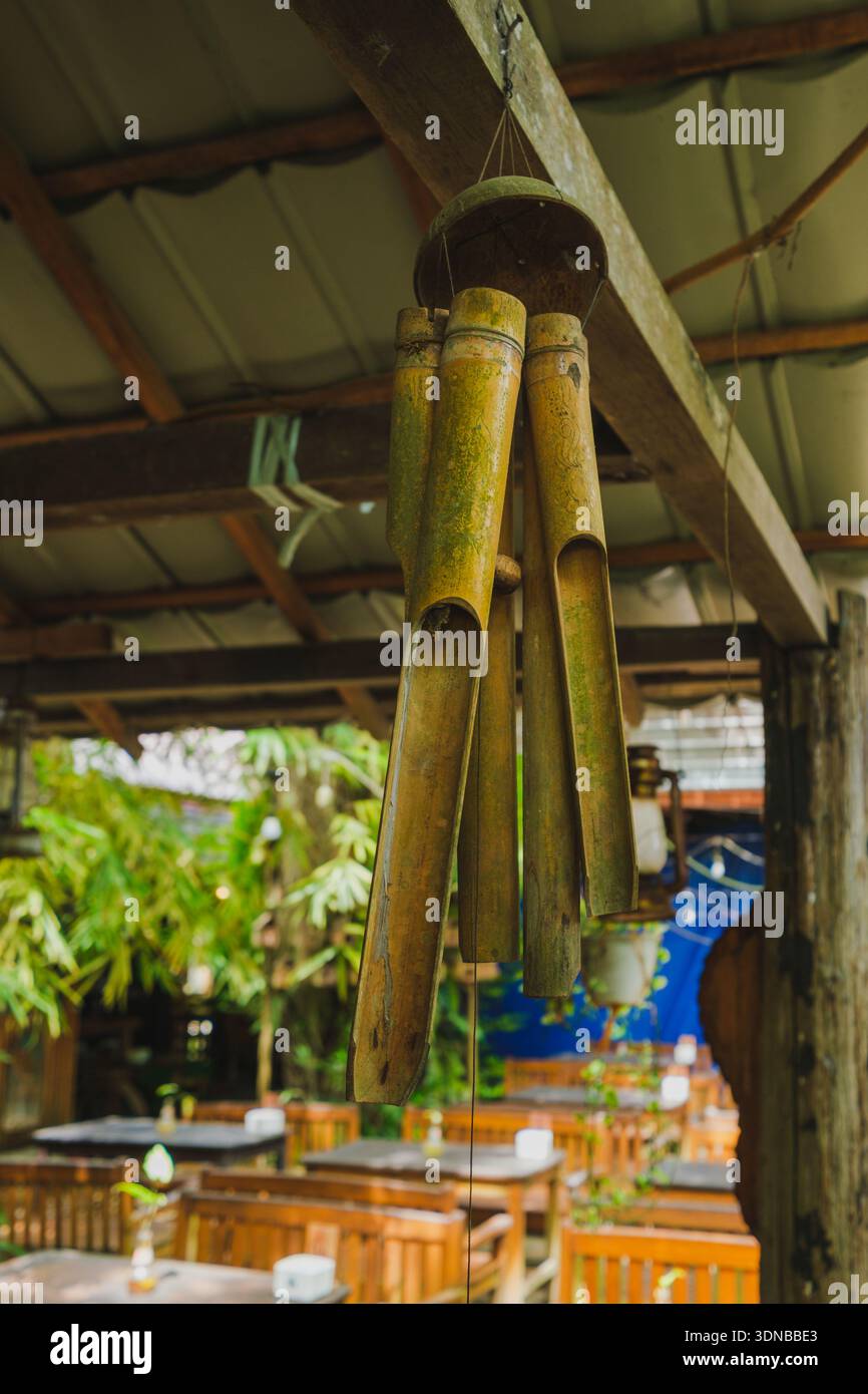 Un primo piano di campanili a vento in bambù fatti a mano (klonthong) appesi a una trave di legno. Lo sfondo rivela una tranquilla e rustica area salotto all'aperto di un Foto Stock