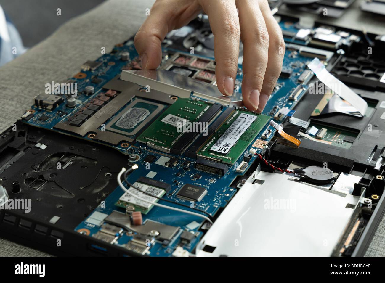Una mano sta sollevando uno schermo metallico da una scheda madre per notebook. L'area di lavoro mostra le parti aperte del laptop e un modulo RAM è pronto per essere installato. Foto Stock